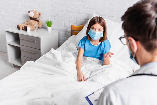 child in medical mask pointing with hand while talking to doctor on blurred foreground