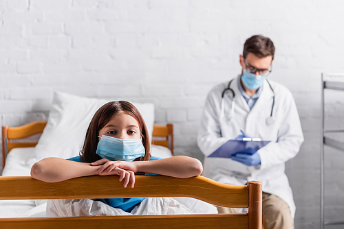 sick girl in medical mask  near doctor writing on clipboard on blurred background
