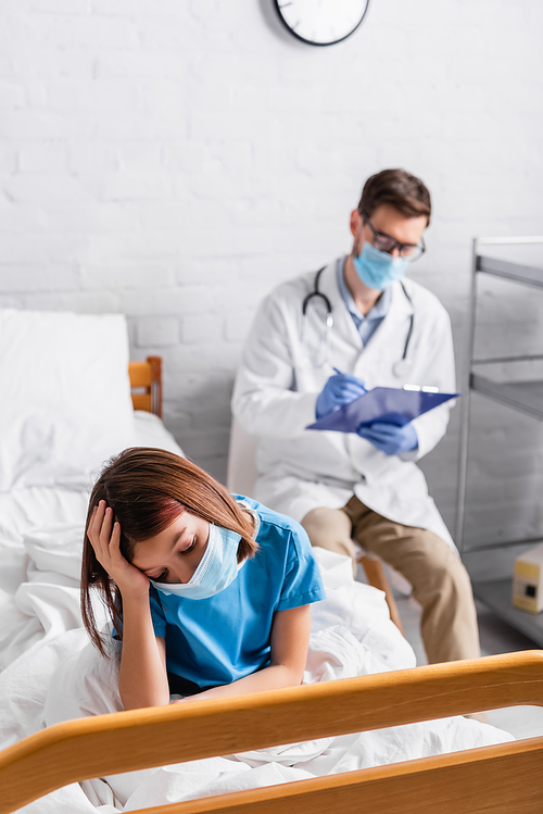 diseased kid in medical mask suffering from headache while doctor writing diagnosis on blurred background
