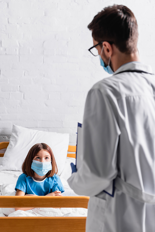 girl in medical mask looking at doctor with clipboard on blurred foreground