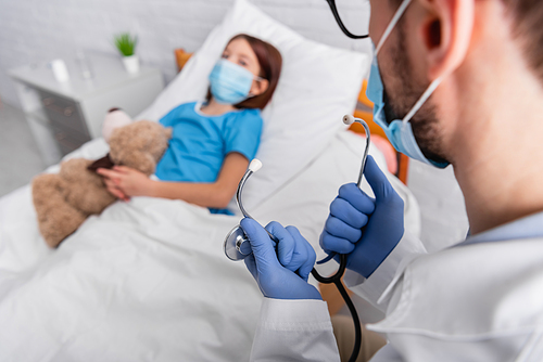 doctor holding stethoscope near sick child in medical mask lying in bed with teddy bear on blurred background