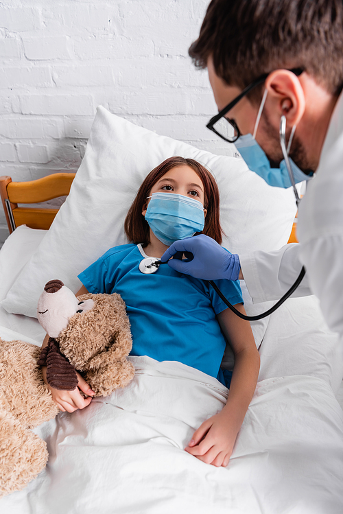 sick girl in medical mask lying in bed with teddy bear while doctor examining girl with stethoscope, blurred foreground