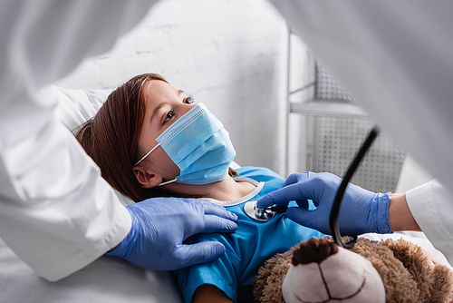 diseased girl in medical mask lying in bed with teddy bear while pediatrician examining her with stethoscope, blurred foreground