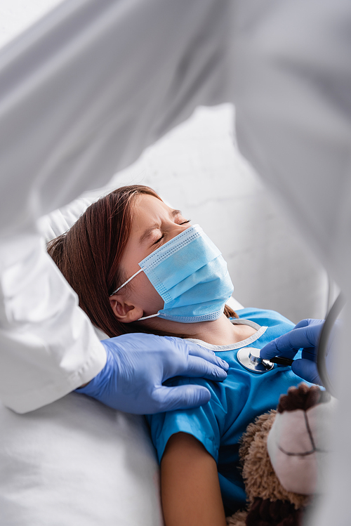 diseased girl in medical mask lying with closed eyes near doctor examining her with stethoscope, blurred foreground