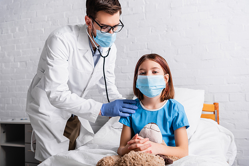 ill girl in medical mask with teddy bear near pediatrician examining her with stethoscope