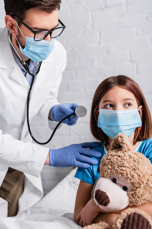 pediatrician with stethoscope near girl in medical mask sitting in bed with teddy bear