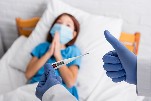 doctor holding thermometer and showing thumb up near child in medical mask lying in bed with praying hands, blurred background