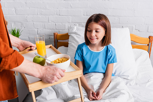 man holding tray with breakfast near daughter in hospital bed, blurred foreground
