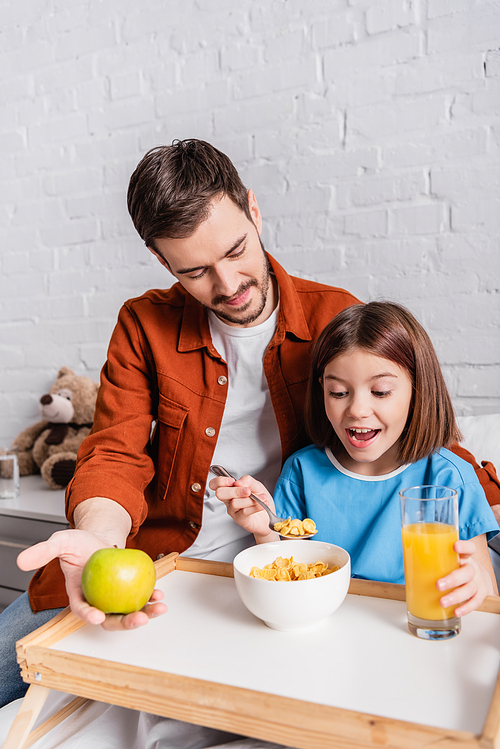 excited girl eating cornflakes near father holding apple in hospital