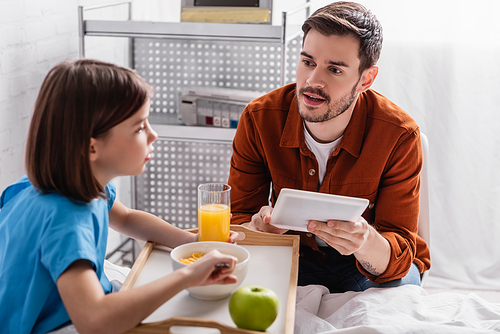 man holding digital tablet near daughter having breakfast in hospital, blurred foreground