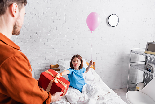 man holding gift box near happy daughter on hospital bed with festive balloon, blurred foreground