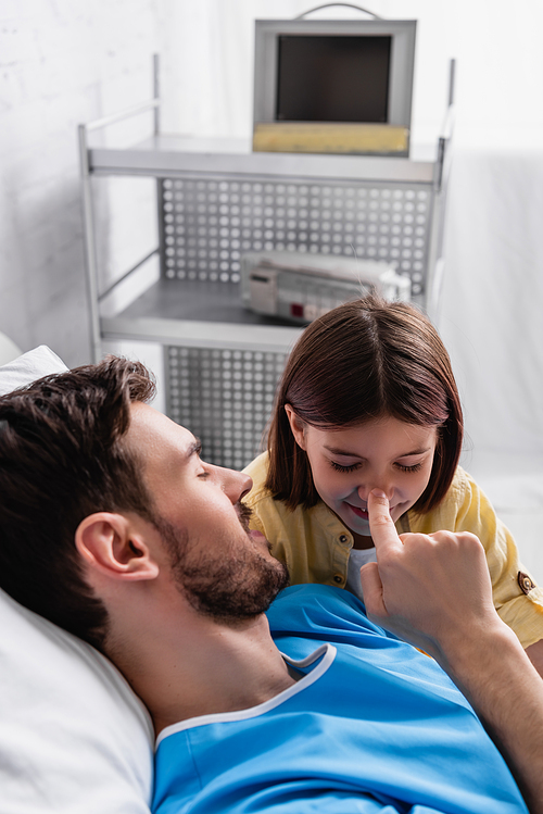 father touching nose of smiling daughter while lying in hospital bed