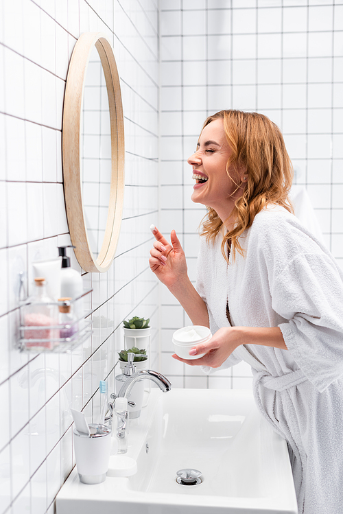 smiling woman holding jar with face cream and laughing near mirror and bottles on blurred foreground