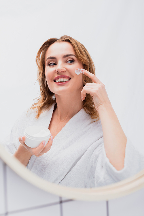 pleased woman holding jar and applying face cream while looking at mirror