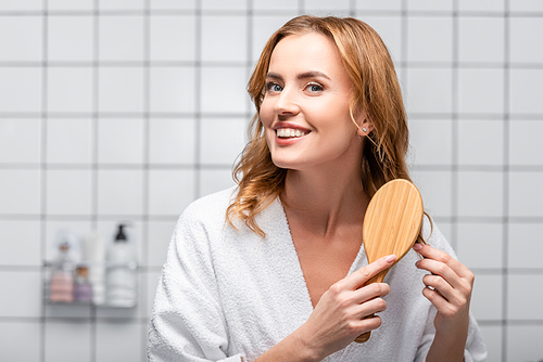 happy woman in white bathrobe brushing hair in bathroom