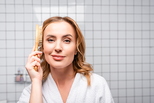 pleased woman in white bathrobe brushing hair in bathroom