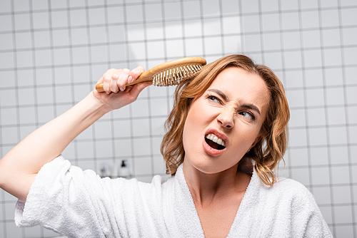 displeased woman in white bathrobe brushing hair in bathroom