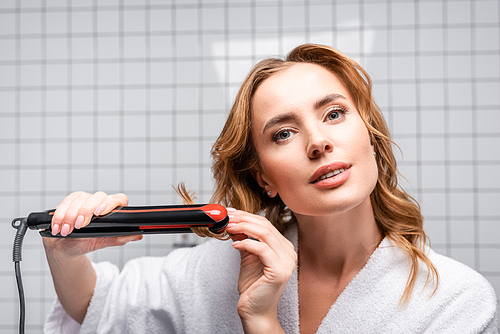 woman in white bathrobe using hair straightener in bathroom