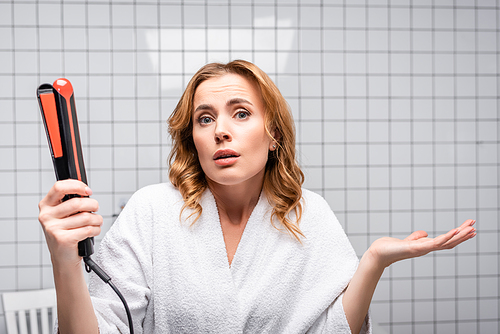 confused woman in white bathrobe holding hair straightener in bathroom