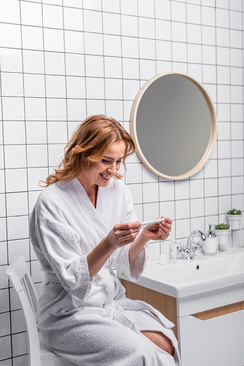 happy woman in white bathrobe looking at pregnancy test in bathroom