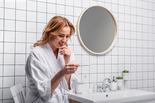 joyful woman in white bathrobe looking at pregnancy test in bathroom