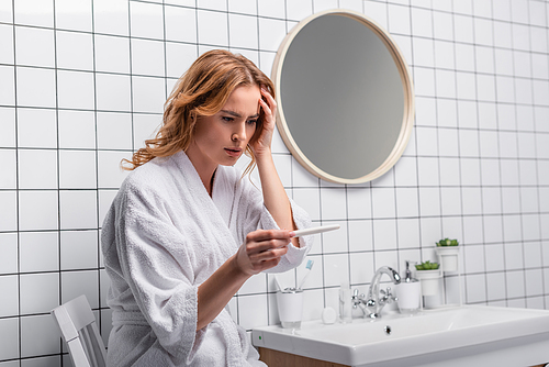 worried woman in white bathrobe holding pregnancy test in bathroom