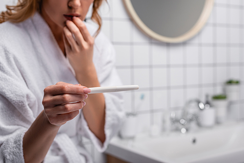 cropped view of worried woman in white bathrobe holding pregnancy test in bathroom