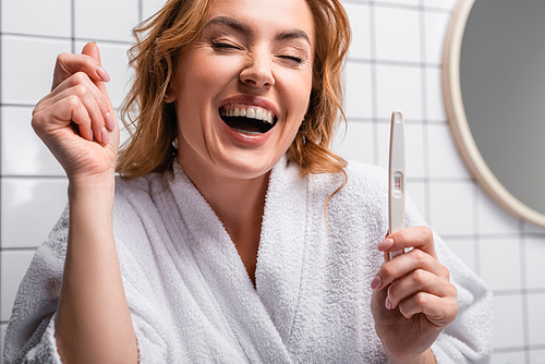 cheerful woman in white bathrobe holding pregnancy test in bathroom