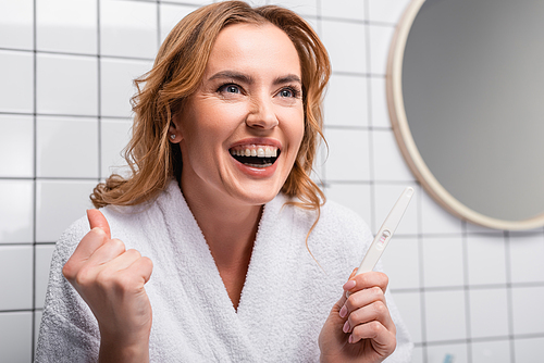 excited woman in white bathrobe holding pregnancy test in bathroom