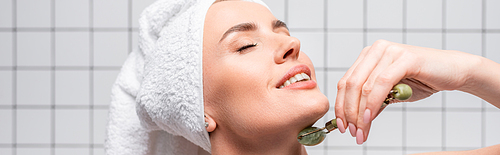 happy woman with closed eyes and towel on head using jade roller in bathroom, banner