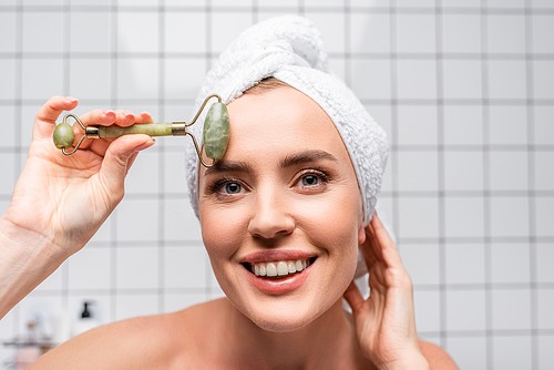 happy woman with towel on head using jade roller in bathroom