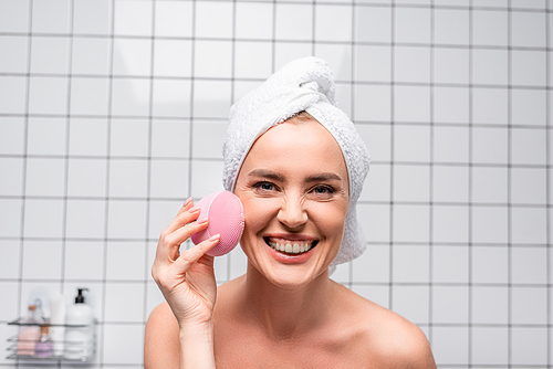 joyful woman in towel on head holding cleansing silicone brush in bathroom