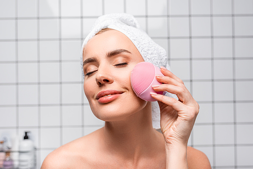 cheerful woman in towel on head and closed eyes holding cleansing silicone brush in bathroom