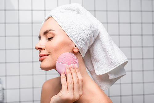 pleased woman in towel on head using cleansing silicone brush in bathroom