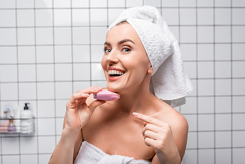 cheerful woman in towel on head pointing with finger at cleansing silicone brush in bathroom