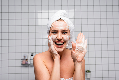 cheerful woman with naked shoulders showing palm with foam cleanser in bathroom