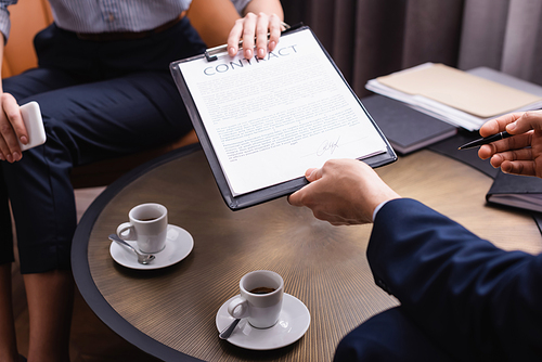 Cropped view of businessman holding contract near businesswoman and coffee on blurred background