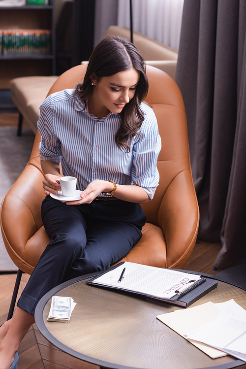 businesswoman with cup of coffee sitting in armchair near contract and dollars on table