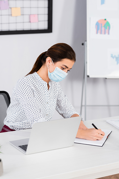 businesswoman in medical mask writing in notebook near laptop