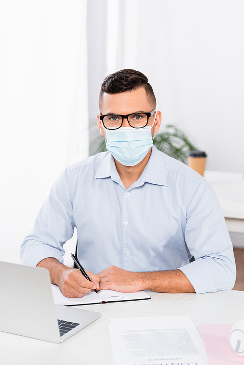 businessman in medical mask and glasses  while holding pen near notebook and laptop on desk