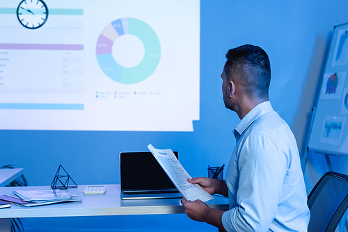 businessman holding contract near laptop with blank screen while looking at charts and graphs on wall