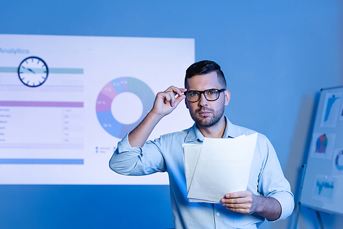businessman adjusting glasses and holding papers near charts and graphs on wall