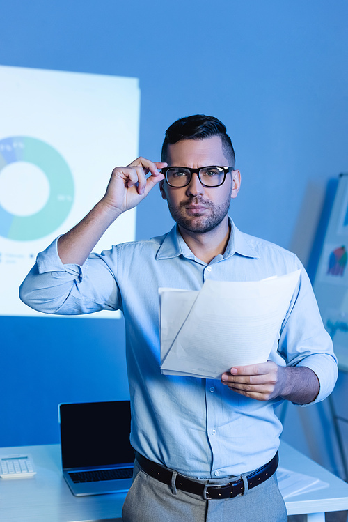 businessman adjusting glasses and holding documents near charts and graphs on wall