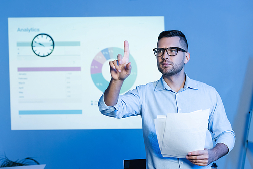businessman in glasses pointing with finger and holding papers near charts on wall