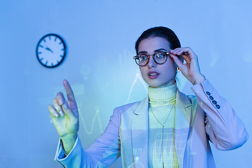 businesswoman in suit adjusting glasses and pointing with finger near digital graphs