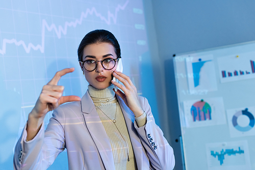 businesswoman in glasses gesturing near digital graphs and talking on smartphone