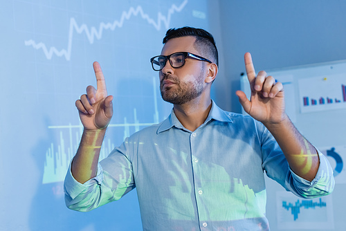 businessman in glasses pointing with fingers near digital graphs in office