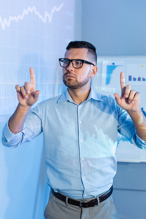 serious businessman in glasses pointing with fingers near digital graphs in office