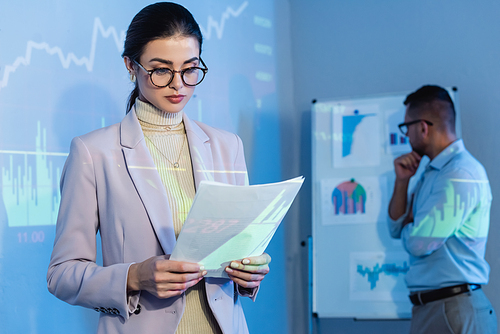 businesswoman in glasses looking at documents near coworker in blurred background