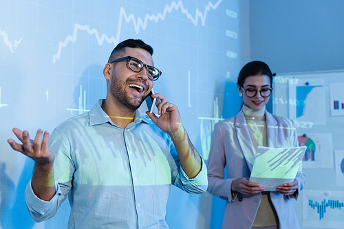 cheerful businessman talking on smartphone near coworker in glasses with documents  on blurred background